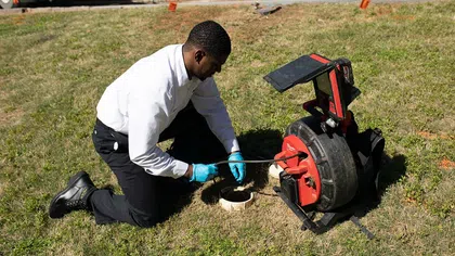 A technician using a plumbing snake at a sewer cleanout on a grassy area.