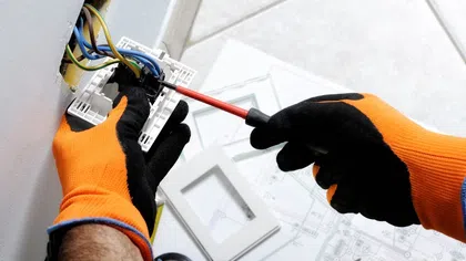 Electrician wearing orange gloves rewiring an electrical outlet.