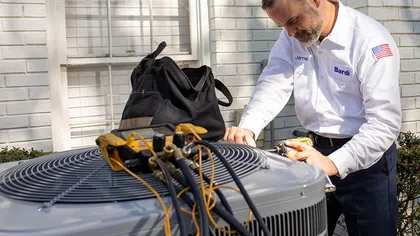 Technician in white uniform repairing an outdoor air conditioning unit with tools and gauges connected.