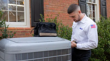 A technician inspects a residential air conditioning unit outside a brick home.