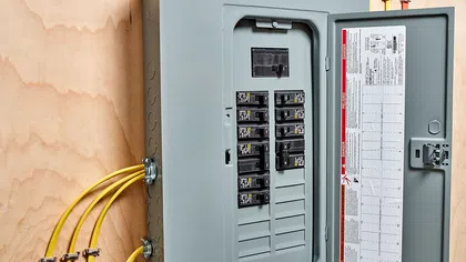 Open electrical panel box with circuit breakers and yellow electrical wires on wooden wall background.
