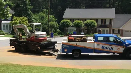 Bardi service truck towing a trailer with a small excavator parked on a residential street near houses.
