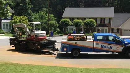 Bardi service truck towing a trailer with a small excavator parked on a residential street near houses.