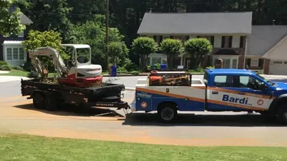 Bardi service truck towing a trailer with a small excavator parked on a residential street near houses.
