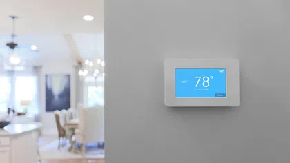 Modern smart thermostat set to 78°F on a gray wall with blurred dining area in the background