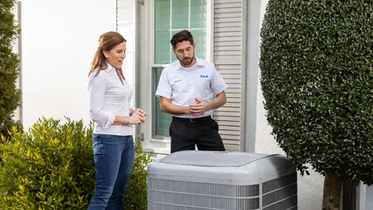 Technician explains air conditioning unit features to homeowner outside a modern house during service appointment.