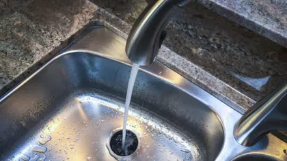 Water flowing from a modern kitchen faucet into a stainless steel sink with droplets on the surface.