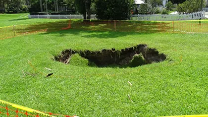 Large sinkhole in a grassy yard surrounded by orange safety fencing near residential houses