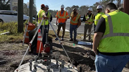 Construction workers in safety vests and helmets inspect concrete foundation with metal bolts by roadside under traffic lights.