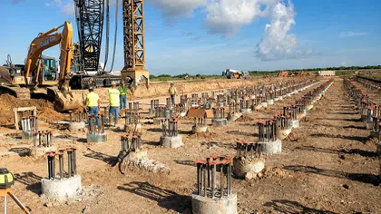 Construction site with concrete foundation pillars and workers operating heavy machinery under a blue sky with clouds.