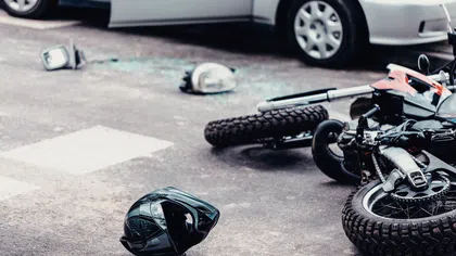 Motorcycle accident scene with fallen bike, helmet, broken headlight, and nearby car on asphalt road.