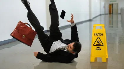 Businessman in suit slipping on a wet floor with briefcase and phone falling beside caution sign in hallway