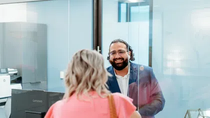 Smiling man in office attire speaking through a clear glass partition to a woman in a pink blouse.