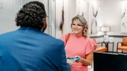 Smiling woman in pink top handing a clipboard to a man in a blue suit at a modern office reception desk.