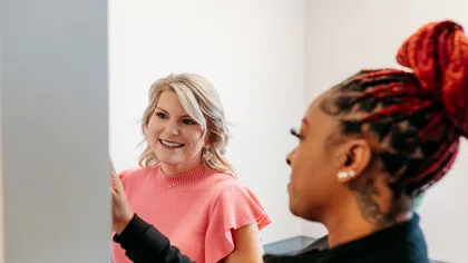 Two women collaborating in an office, one pointing at a screen and the other attentively smiling and listening.