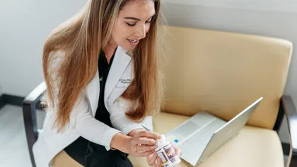 Woman in white coat sitting on beige couch holding a medicine bottle with a laptop open beside her.
