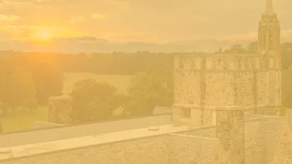 Stone building with tower at sunset overlooking green fields and distant mountains under a cloudy sky