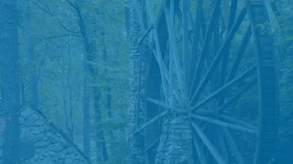 Historic wooden waterwheel attached to a stone mill building surrounded by lush green forest trees.
