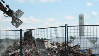 Construction machinery demolishing a building with debris behind a safety fence under a partly cloudy sky.