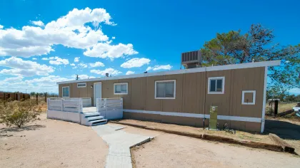 Single-story beige mobile home with white trim and small porch under a bright blue sky with scattered clouds.