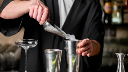 Bartender pouring ice cubes into a metal cocktail shaker with an empty glass and jigger on a bar counter.