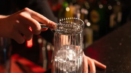 Bartender uses a cocktail strainer over a glass filled with ice on a dimly lit bar counter.