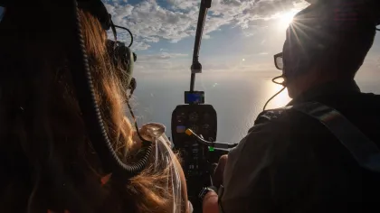 Two people piloting a helicopter over the ocean with the sun shining through partly cloudy sky.