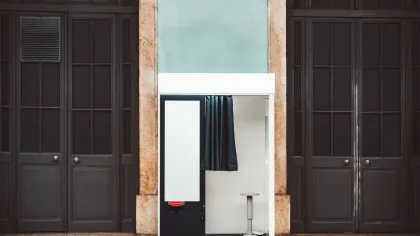 Small photo booth with black curtain and stool against stone and teal walls between two dark wooden doors.