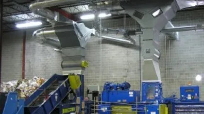 Industrial recycling facility with conveyor belt, baler machine, and stacks of compressed cardboard inside warehouse.