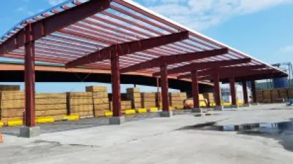 Open warehouse with red steel roof structure storing stacks of lumber under clear sky.
