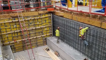 Two construction workers in safety gear working on concrete wall reinforcement at a large building site.