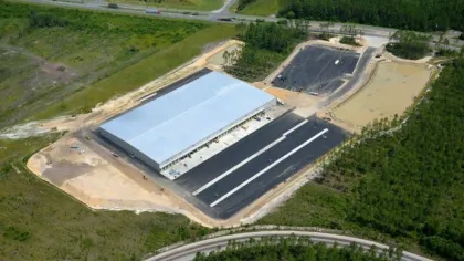 Aerial view of a large warehouse with adjacent paved loading areas surrounded by greenery and roads.