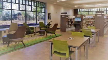 Modern library interior with green chairs, study tables, lounge seating, large windows, and bookshelves.