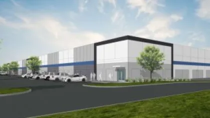 Modern industrial warehouse building with parking lot, parked cars, and landscaped greenery under a partly cloudy sky