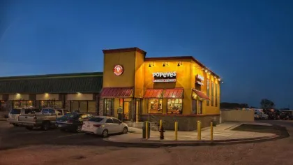 Popeyes Louisiana Kitchen fast food restaurant illuminated at dusk with cars parked outside and dark blue sky.