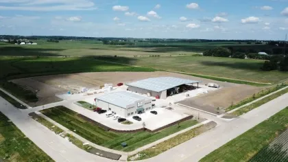Aerial view of a modern industrial building and warehouse surrounded by farmland and roads under a blue sky.