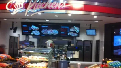 Convenience store kitchen area with food displays, menu screens, and snack aisles under Kitchen's sign