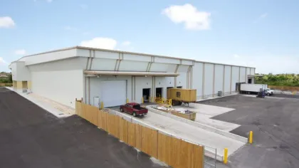 Modern warehouse building with loading docks, trucks, and a fenced perimeter under a clear blue sky.