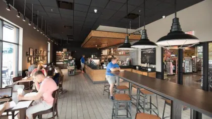 Modern coffee shop interior with wooden accents, pendant lights, people working and socializing, and large windows.