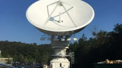 Large white satellite dish antenna positioned outdoors with trees and equipment under clear blue sky.