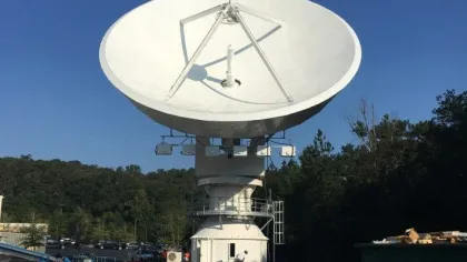 Large white satellite dish antenna outdoors with clear blue sky and surrounding trees in the background.