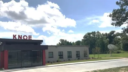 Modern building with KNOE sign on rooftop, satellite dish, trees, and clear blue sky with clouds.