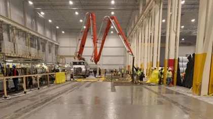 Workers using concrete pumps inside a large industrial warehouse under bright lights.