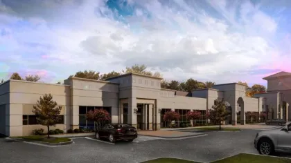 Modern commercial office building with parking lot, trees, and a partly cloudy blue sky at dusk.