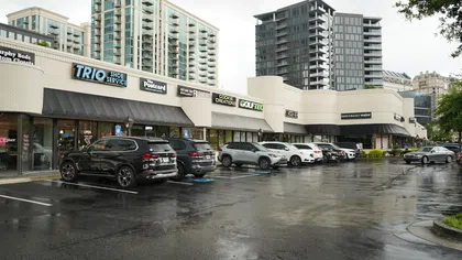 Shopping center with parked cars on wet pavement under cloudy sky in urban area with high-rise buildings.