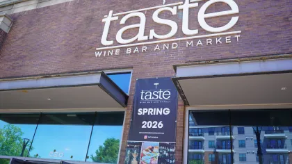 Taste Wine Bar and Market storefront with a Spring 2026 event banner on a brick building under blue sky.