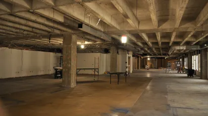 Large empty industrial space under renovation with concrete pillars, exposed ceiling, and construction equipment.