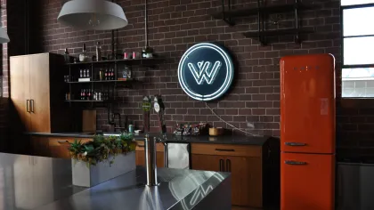 Industrial style kitchen with brick walls, neon sign, orange retro fridge, wooden cabinets, and modern stainless steel island.