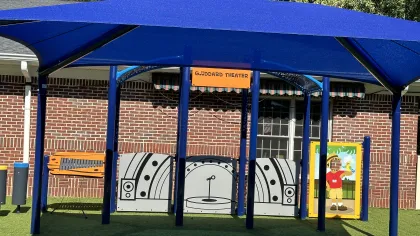 Outdoor children's play theater area with blue canopy, stage backdrop, and artificial grass seating resembling rocks.