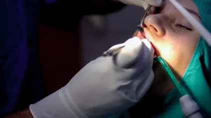 Dentist wearing gloves treating a patient's teeth with dental tools under focused light during a procedure.